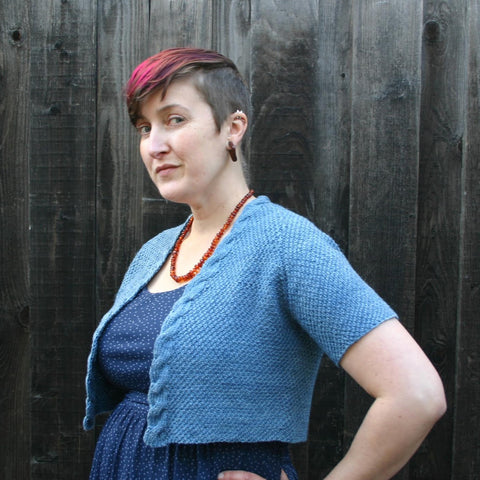 A white woman with close cropped brown hair that is dyed magenta on top is standing in front of a wooden fence.  She is standing at quarter profile with her hand on her hip.  She is wearing a blue dress with a blue, cropped, textured cardigan over it and an amber bead necklace. 